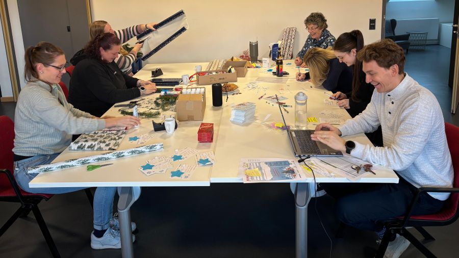 Sieben Menschen sitzen an einem großen Tisch und helfen beim Geschenkeverpacken.