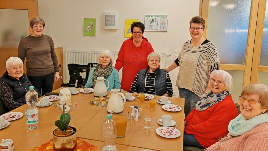 Das Foto zeigt eine Gruppe Menschen, teilweise sitzend, teilweise stehend um einen gedeckten Tisch.