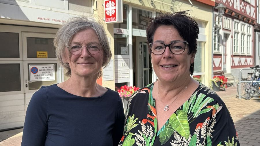 Zwei Frauen stehen vor einem Hauseingang, leicht verschwommen ist das Caritas-Logo am Haus erkennbar.