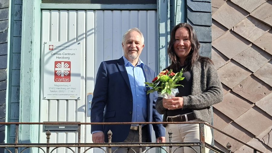 Zwei Menschen vor einer Tür mit Caritas-Flamme, die Frau hält einen Blumenstrauß.