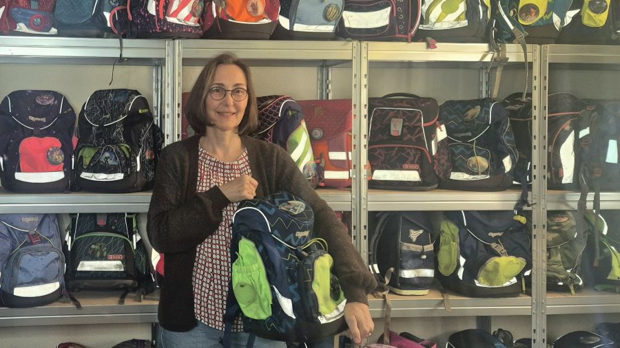 Eine Frau zeigt Schultaschen, hinter ihr ein Regal voller Schulranzen.