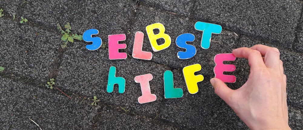 Schriftzug Selbsthilfe als Foto: eine Hand legt die Buchstaben