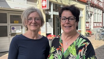 Zwei Frauen stehen vor einem Hauseingang, leicht verschwommen ist das Caritas-Logo am Haus erkennbar.