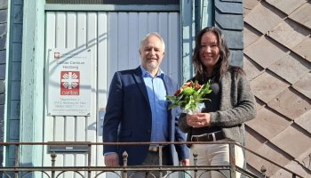 Zwei Menschen vor einer Tür mit Caritas-Flamme, die Frau hält einen Blumenstrauß.