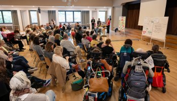 Menschen sitzen im Halbkreis, einige im Vordergrund auf eigenen Rollstühlen.