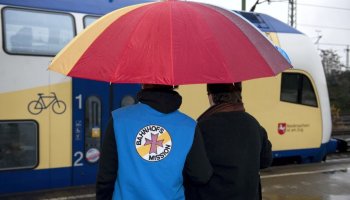Das Foto zeigt zwei Personen unter einem Regenschirm auf dem Weg zum Zug, der im Hintergrund steht.