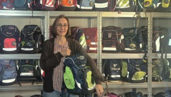 Eine Frau zeigt Schultaschen, hinter ihr ein Regal voller Schulranzen.