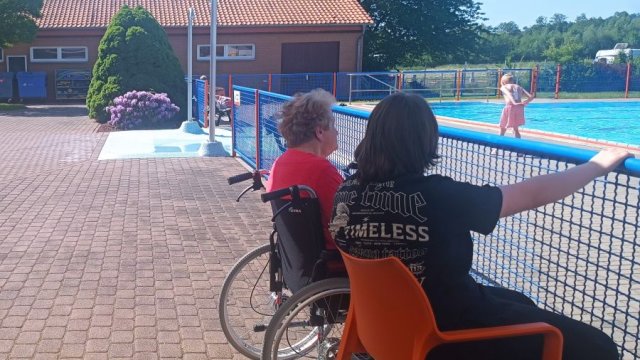 Das Foto zeigt zwei Personen, davon eine im Rollstuhl, am Wasserrand des Freibads.