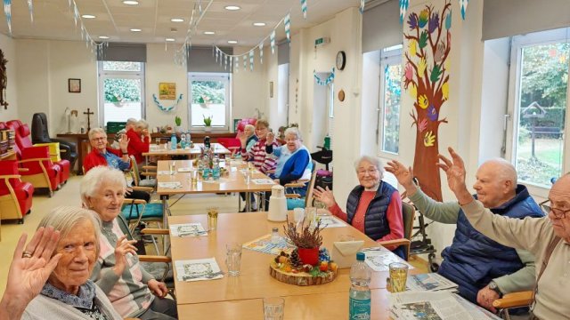 Die Menschen sitzen an einer langen Tafel und winken in die Kamera.