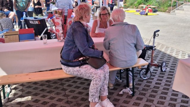 Im Vordergrund sitzen drei Personen an einer Biertisch-Garnitur.