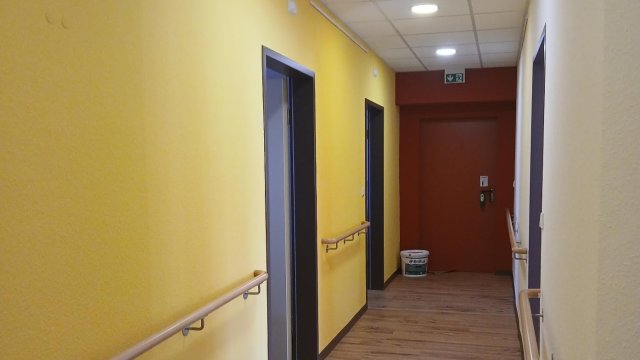 Das Foto zeigt einen Flur mit einer gelben Wand links, mit einer roten Fahrstuhltür geradeaus und mit einer weißen Wand rechts.