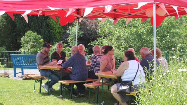 Das Foto zeigt eine Gruppe Menschen, die unter einem roten Pavillon an einer Birbank sitzen.