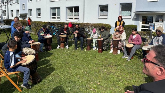 Das Foto zeigt eine Gruppe Menschen, die im Kreis sitzen und Trommeln zwischen ihren Beinen halten.